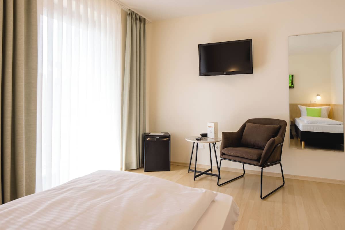Una habitación de hotel con cama, una silla, televisor, mesita y espejo.