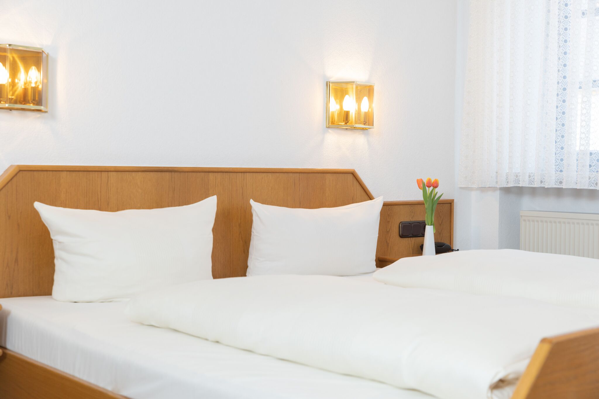 Una cama bien hecha con sábanas blancas, cabecera de madera y dos lámparas fijadas a la pared.