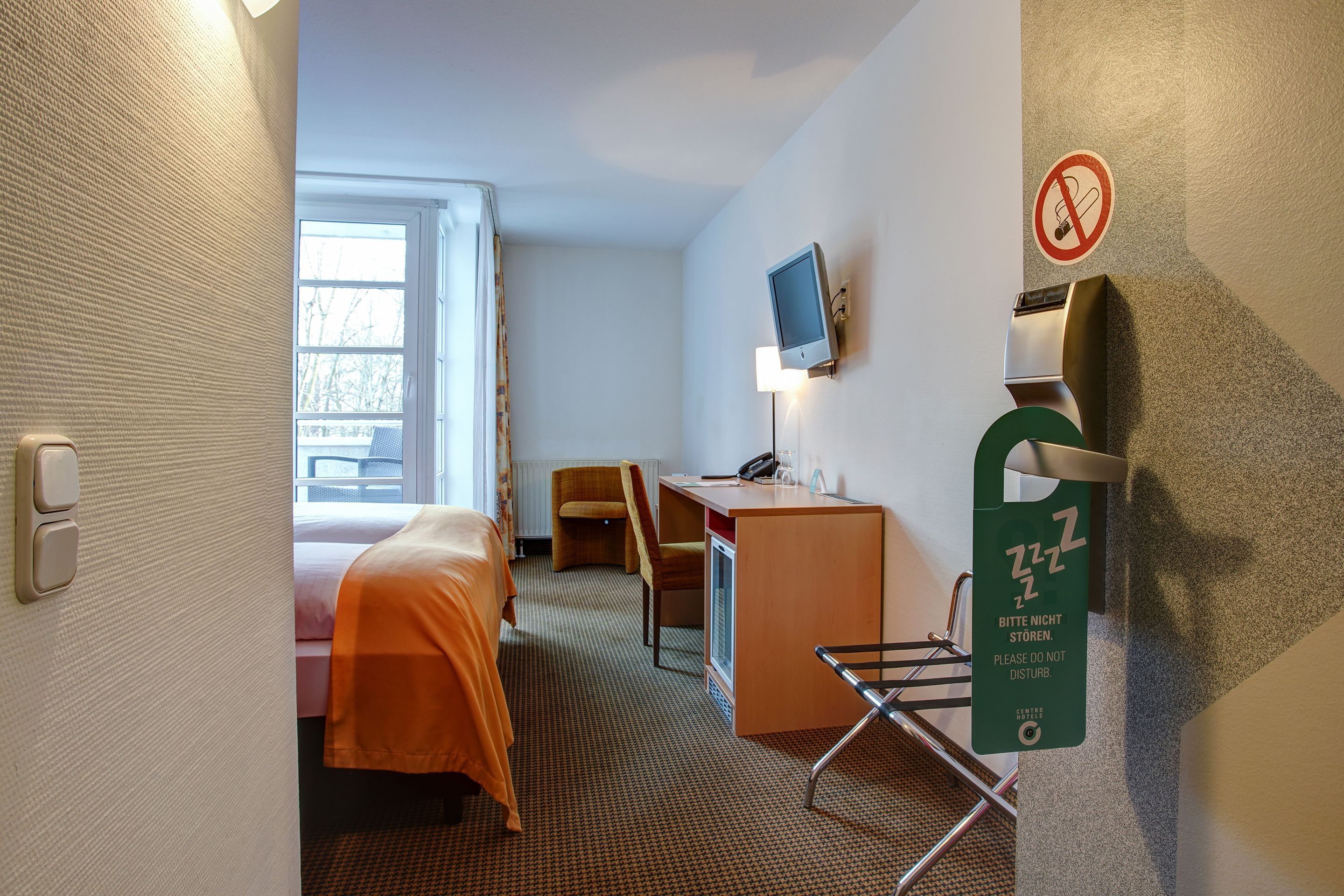 A hotel room with a bed, desk, chair, TV, and a sign indicating quiet hours.