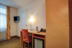 Una habitación de hotel con escritorio, silla, televisor, lámpara, teléfono y un refrigerador pequeño.