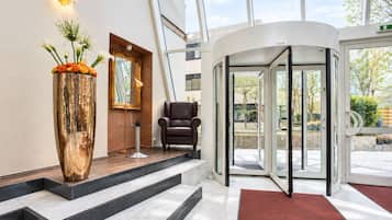Eine Hotelhalle mit einer Kreiseltür, einem Ledersessel, einer großen Vase mit Blumen und einem Spiegel an der Wand.
