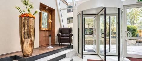 A hotel lobby with a revolving door, a leather chair, a large vase with flowers, and a mirror on the wall.