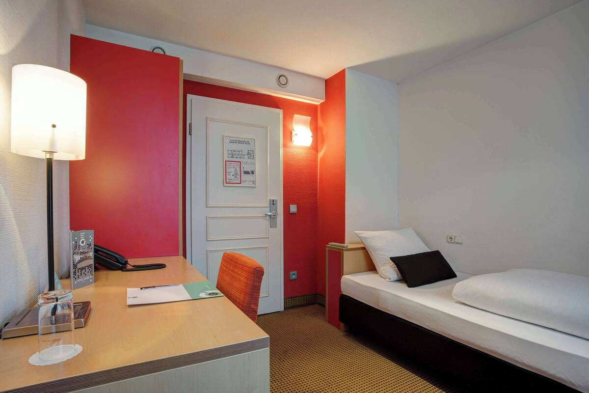 A hotel room with a desk, a bed, a chair, a lamp, and a wall-mounted light.