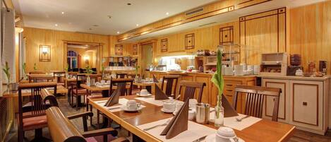 A hotel dining area with wooden furniture, multiple dining tables, and a kitchen in the background.