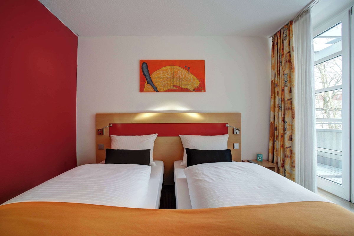 Habitación de hotel con dos camas, una pared de acento roja, un cuadro y una ventana con cortinas.