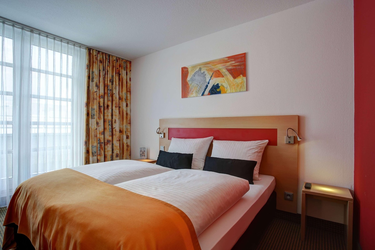 Habitación de hotel con una cama grande, cabecero, un cuadro en la pared y una ventana con cortinas.