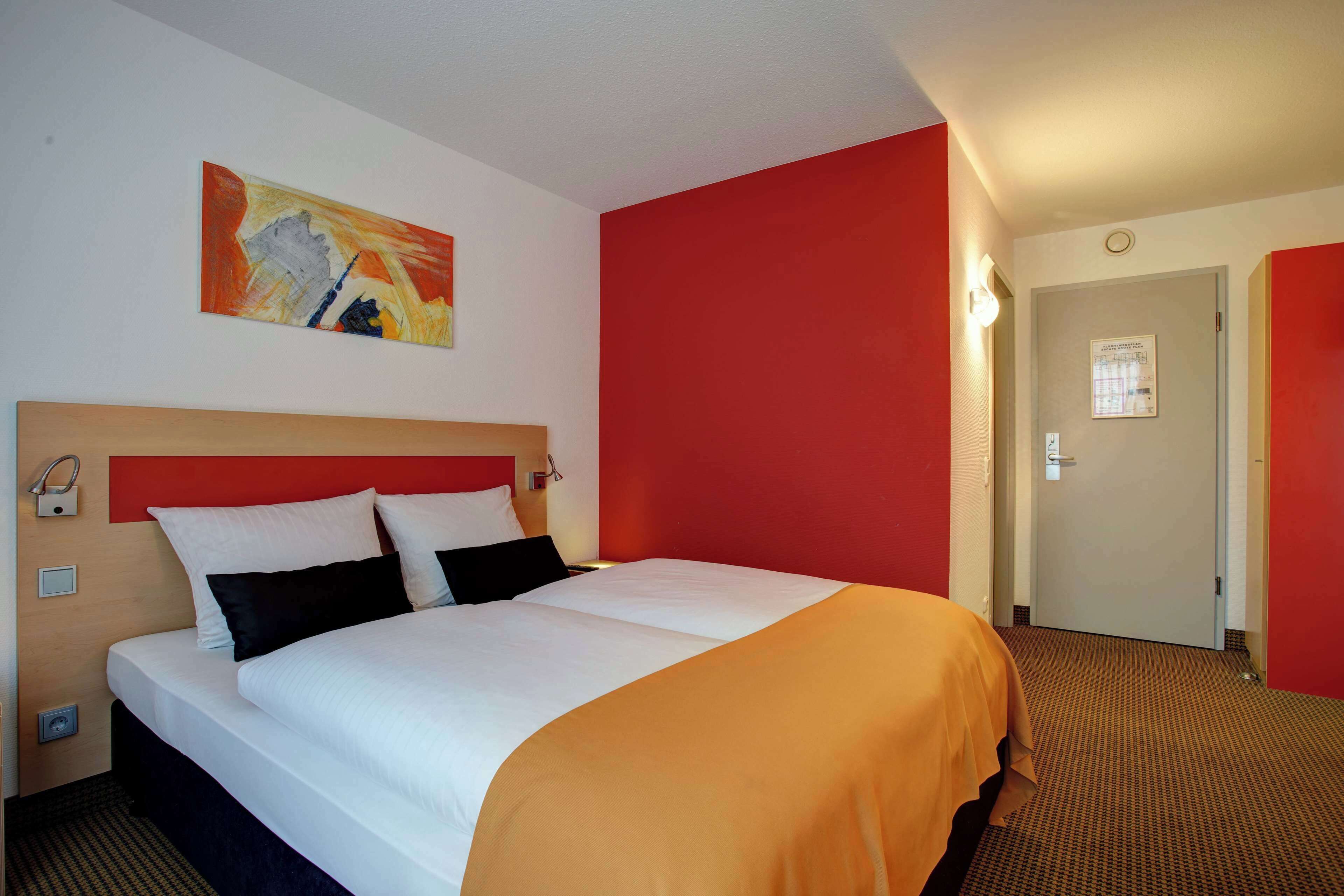 A hotel room with a large bed, a red accent wall, and a painting above the bed.