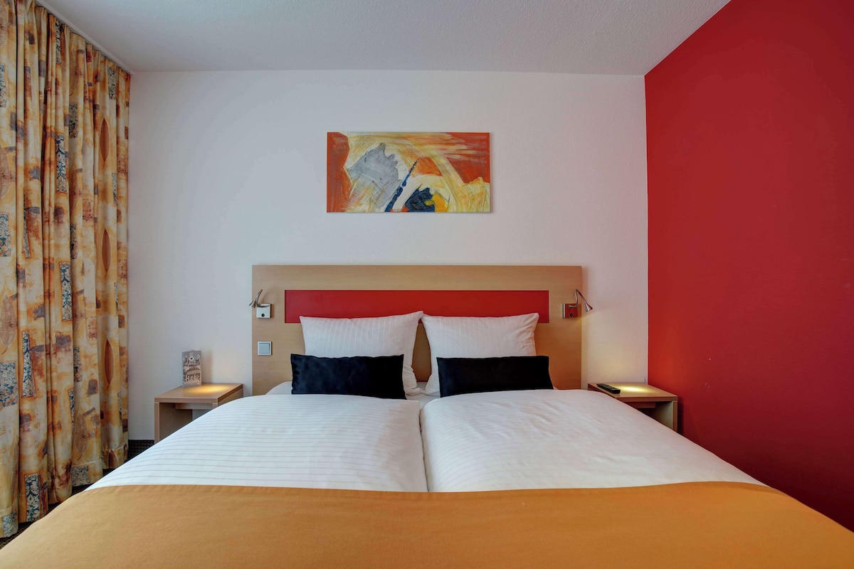 Una habitación de hotel con una cama, dos mesitas de noche, una pared de acento roja y un cuadro sobre la cama.