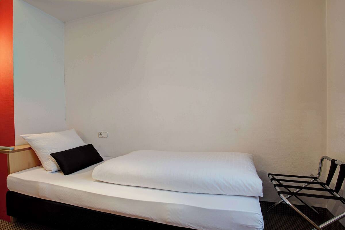A single bed with white bedding and a black pillow, a metal chair, and a red accent wall.