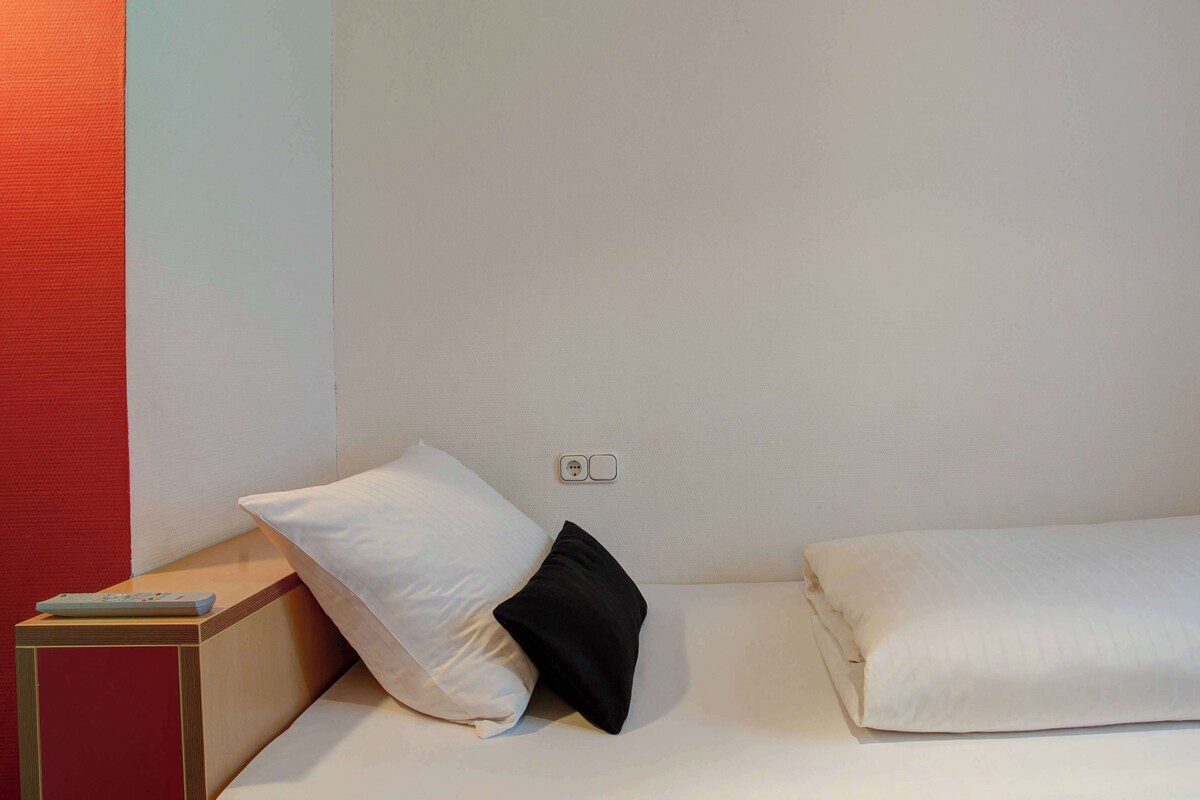 Una cama individual con ropa de cama blanca y una almohada negra, una mesita de noche de madera con un control remoto y un enchufe de pared.