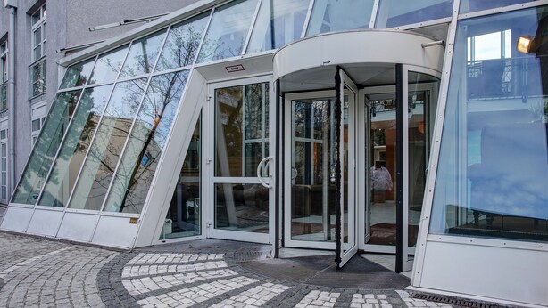 Una entrada de edificio moderna con fachada de vidrio, puerta de cristal curva y zona de acceso con baldosas.