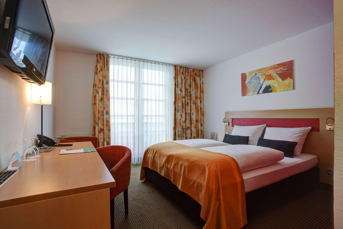 Habitación de hotel con cama, escritorio, silla, televisor y ventana con cortinas.