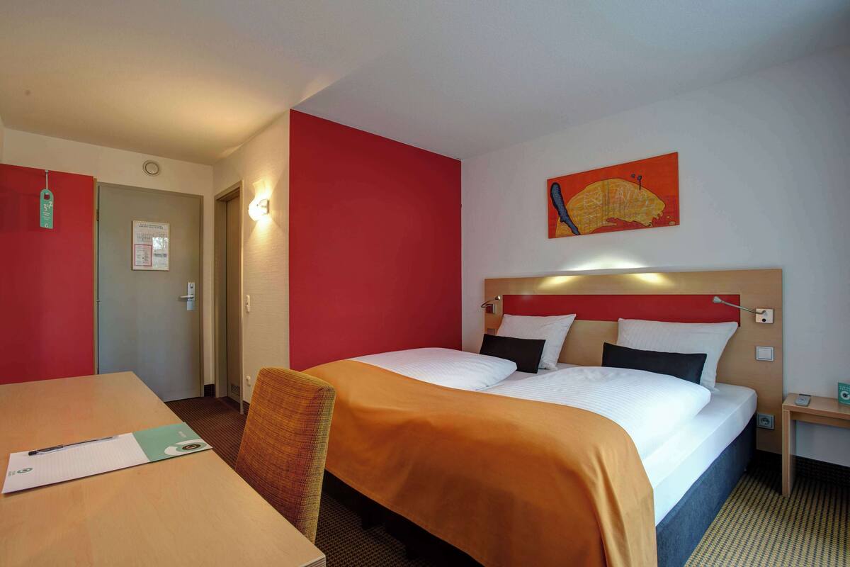 A hotel room with a bed, a desk, and a chair. There is a painting on the wall and a door to the left.