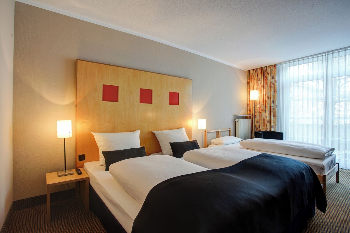 Habitación de hotel con dos camas, cabecero de madera, lámparas de noche y un ventanal con cortinas.