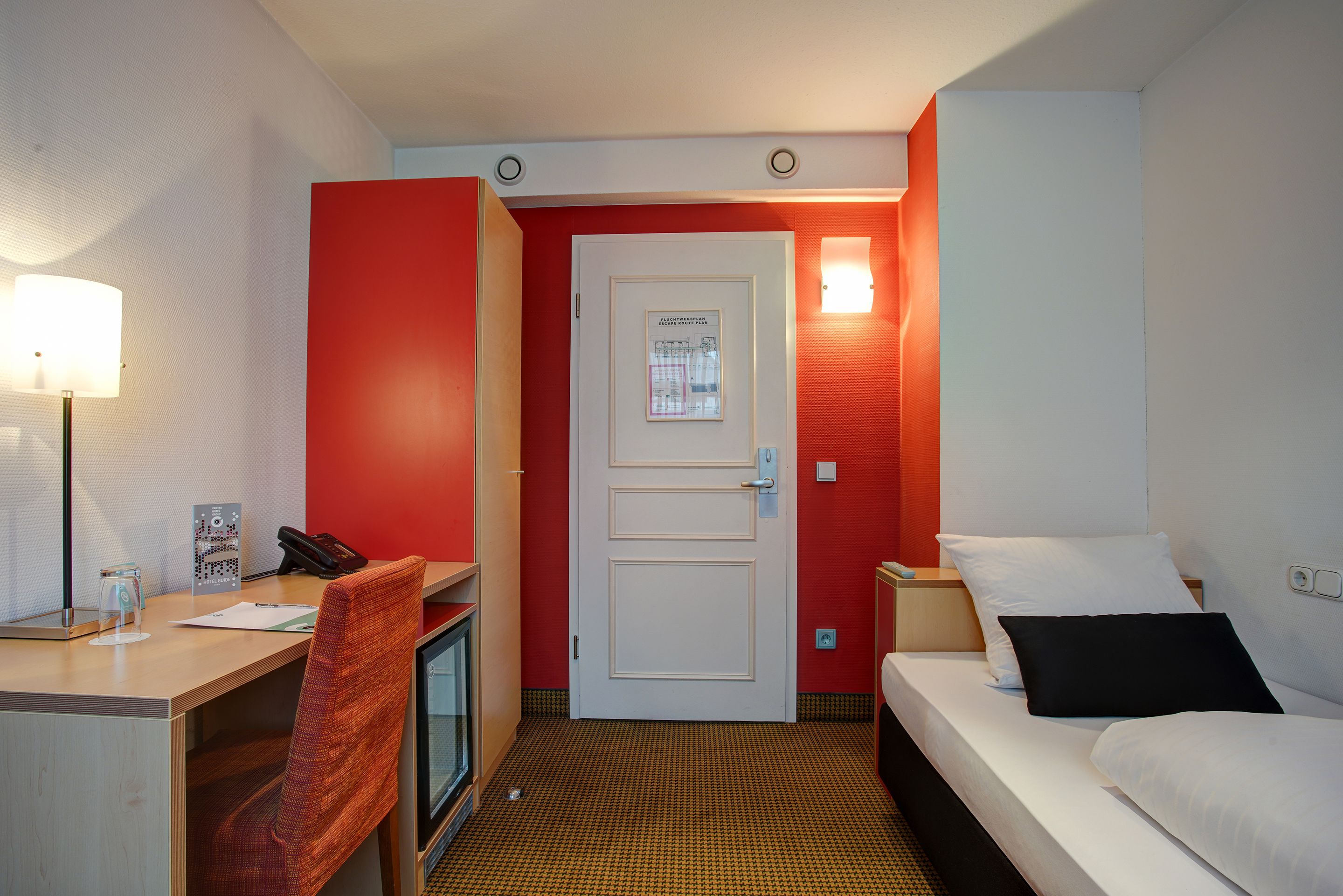 A hotel room with a desk, chair, bed, and a door with a poster.