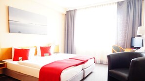 Habitación de hotel con cama, una silla, escritorio con lámpara y un gran ventanal con cortinas.