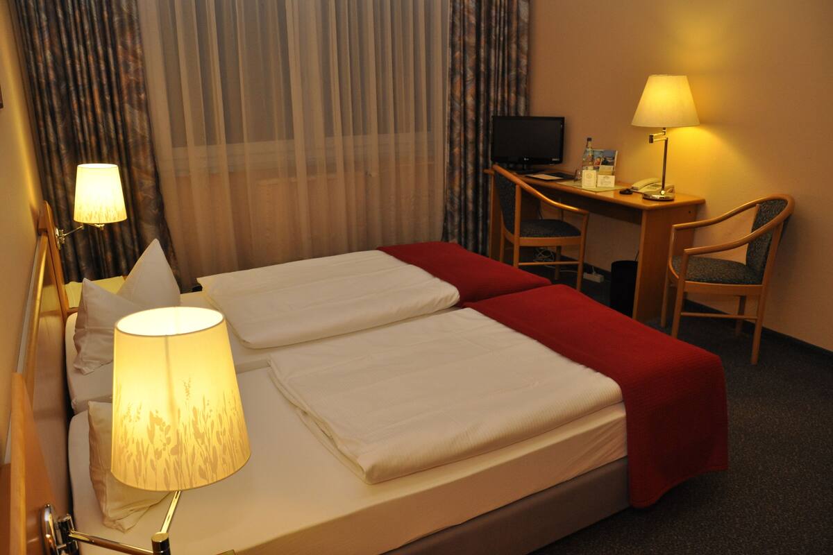 A hotel room with a bed, two bedside lamps, a desk with a computer, and a chair.
