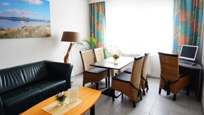 Una habitación de hotel con un sofá de cuero negro, una mesa de comedor con sillas de mimbre, una computadora portátil sobre un escritorio y un cuadro de un faro en la pared.