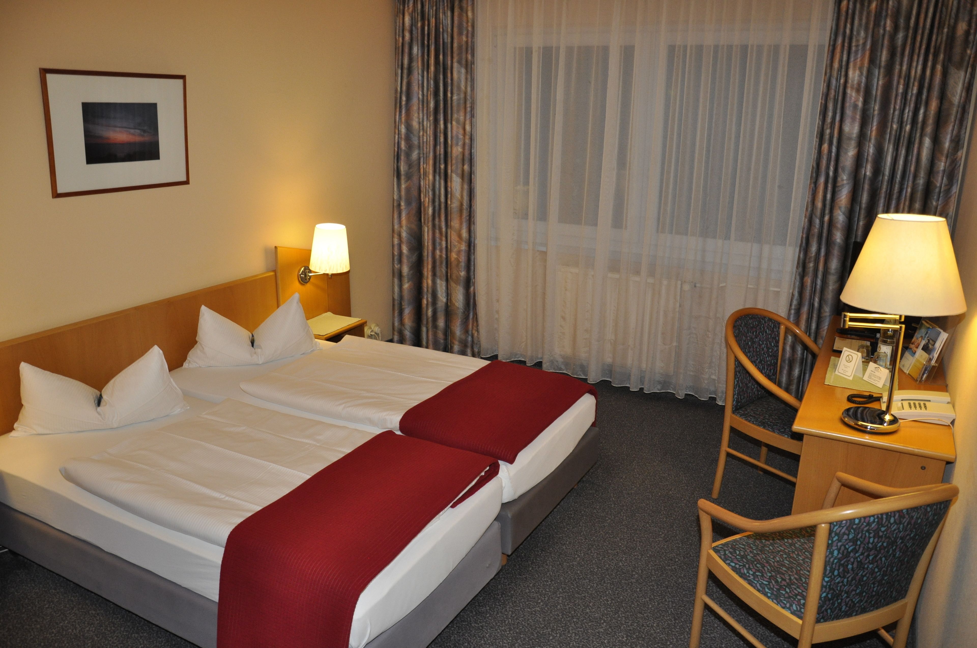 Een hotelkamer met een tweepersoonsbed, twee nachtkastjes met lampen, een bureau met stoel en een raam met gordijnen.