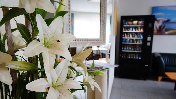 Una habitación con un ventanal grande, una máquina expendedora negra y una mesa con un jarrón de lirios blancos.