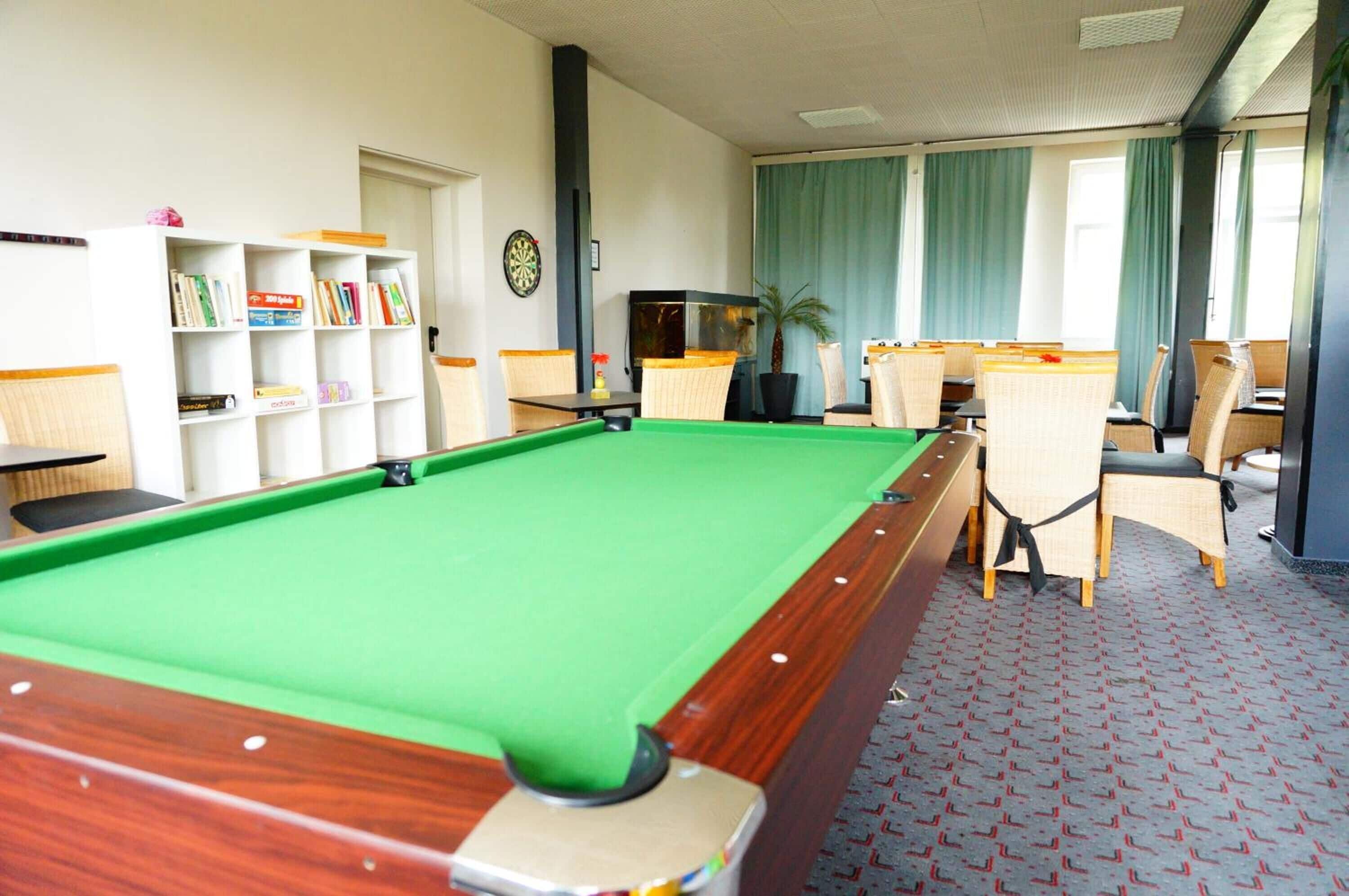 A pool table with a green felt surface, surrounded by chairs and a bookshelf with books.