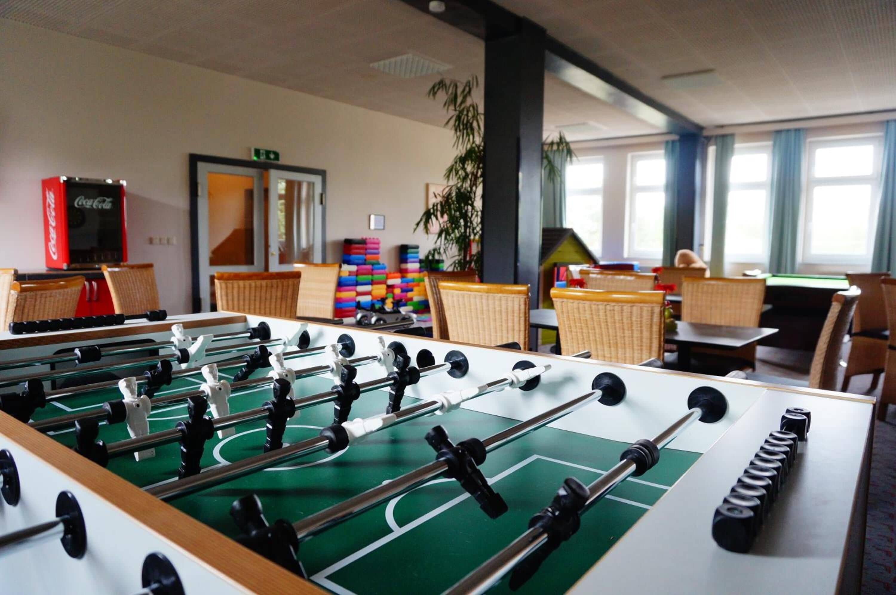A foosball table in a room with a Coca-Cola refrigerator, a door with a mirror, and a plant.
