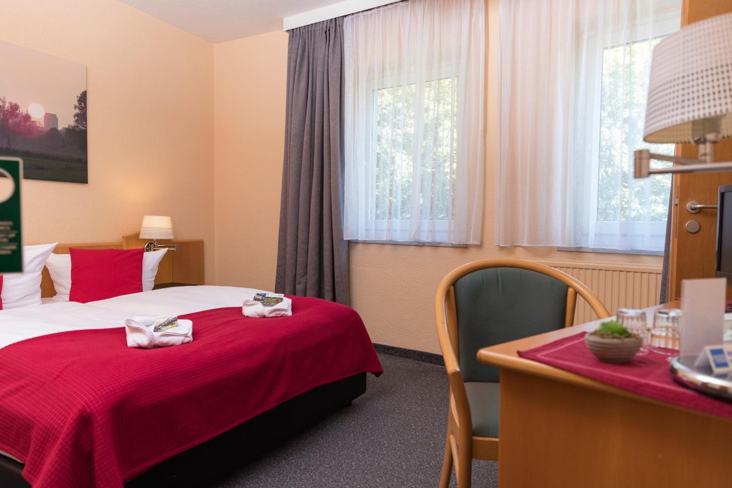 Habitación de hotel con cama, escritorio, silla, ventana con cortinas y lámpara.