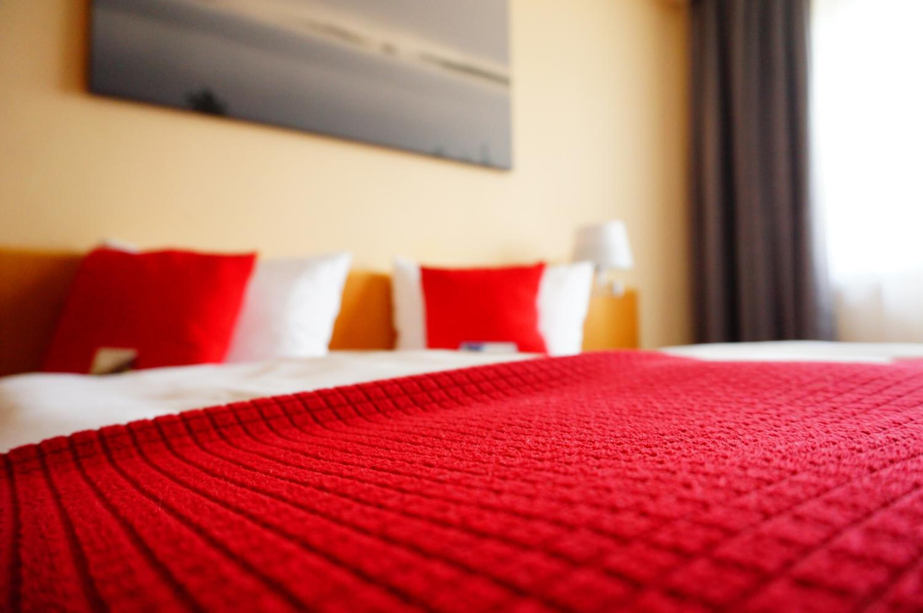 A bed with a red blanket and pillows, a white headboard, and a painting on the wall.