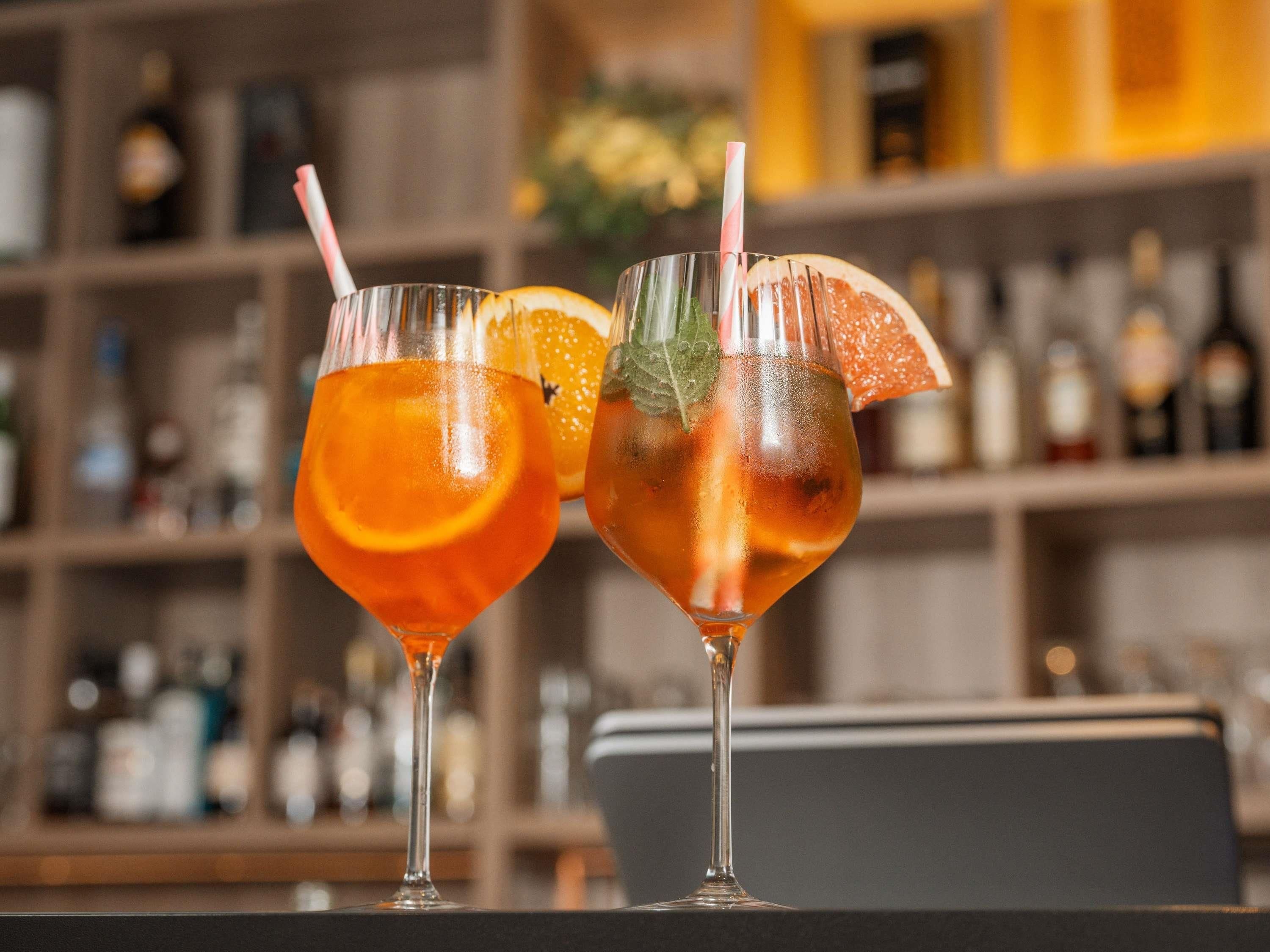 To cocktails i høje glas med pynt, placeret på en bardisk med hylder fyldt med flasker i baggrunden.