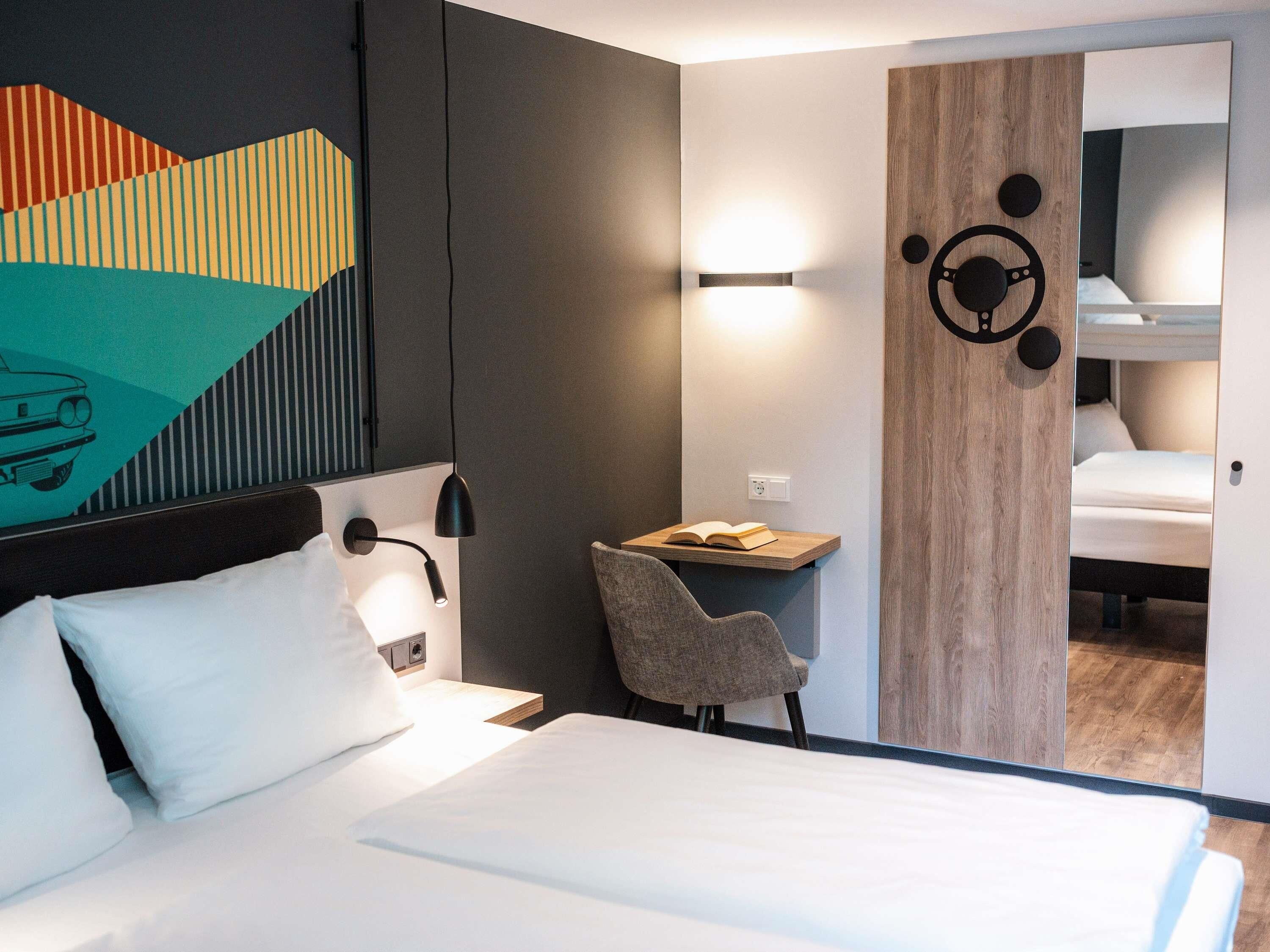 Une chambre d’hôtel comprenant un lit, un bureau avec une chaise, un luminaire fixé au mur et une porte avec un panneau en bois.