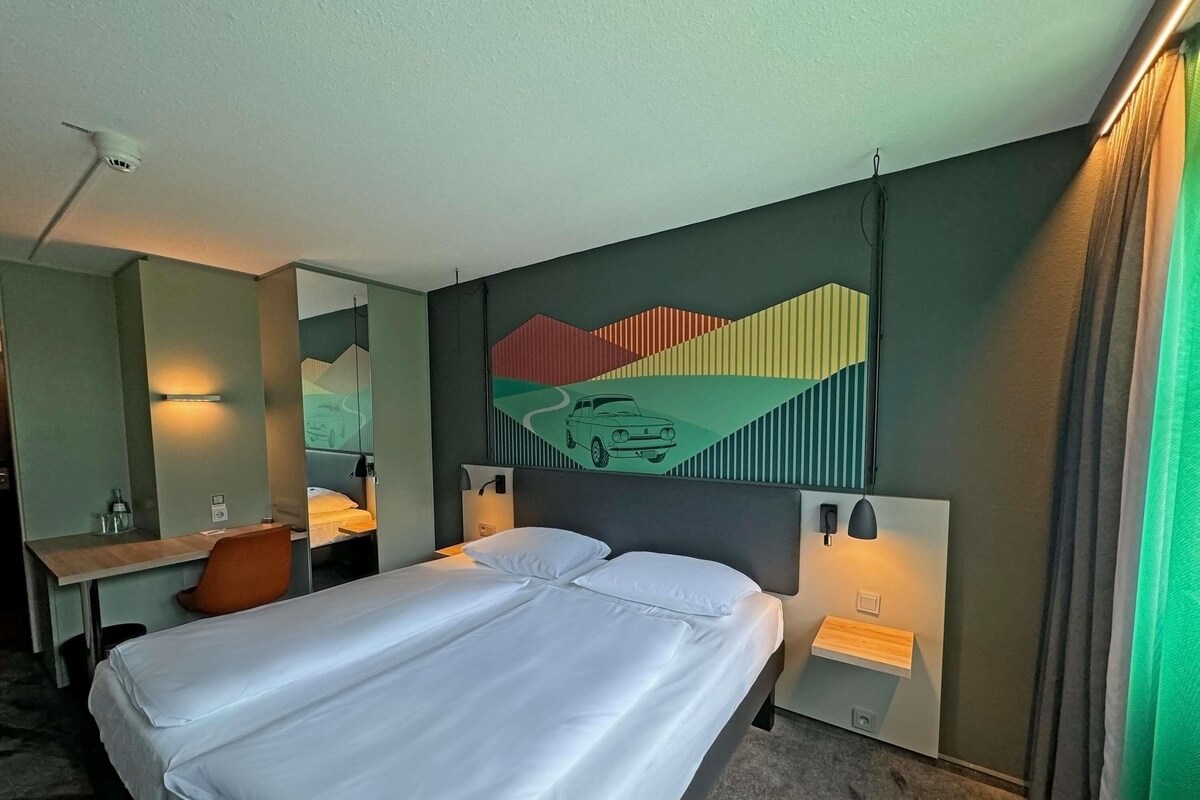 A hotel room with a large bed, a desk with a chair, a mirror, and a wall with a colorful abstract painting.