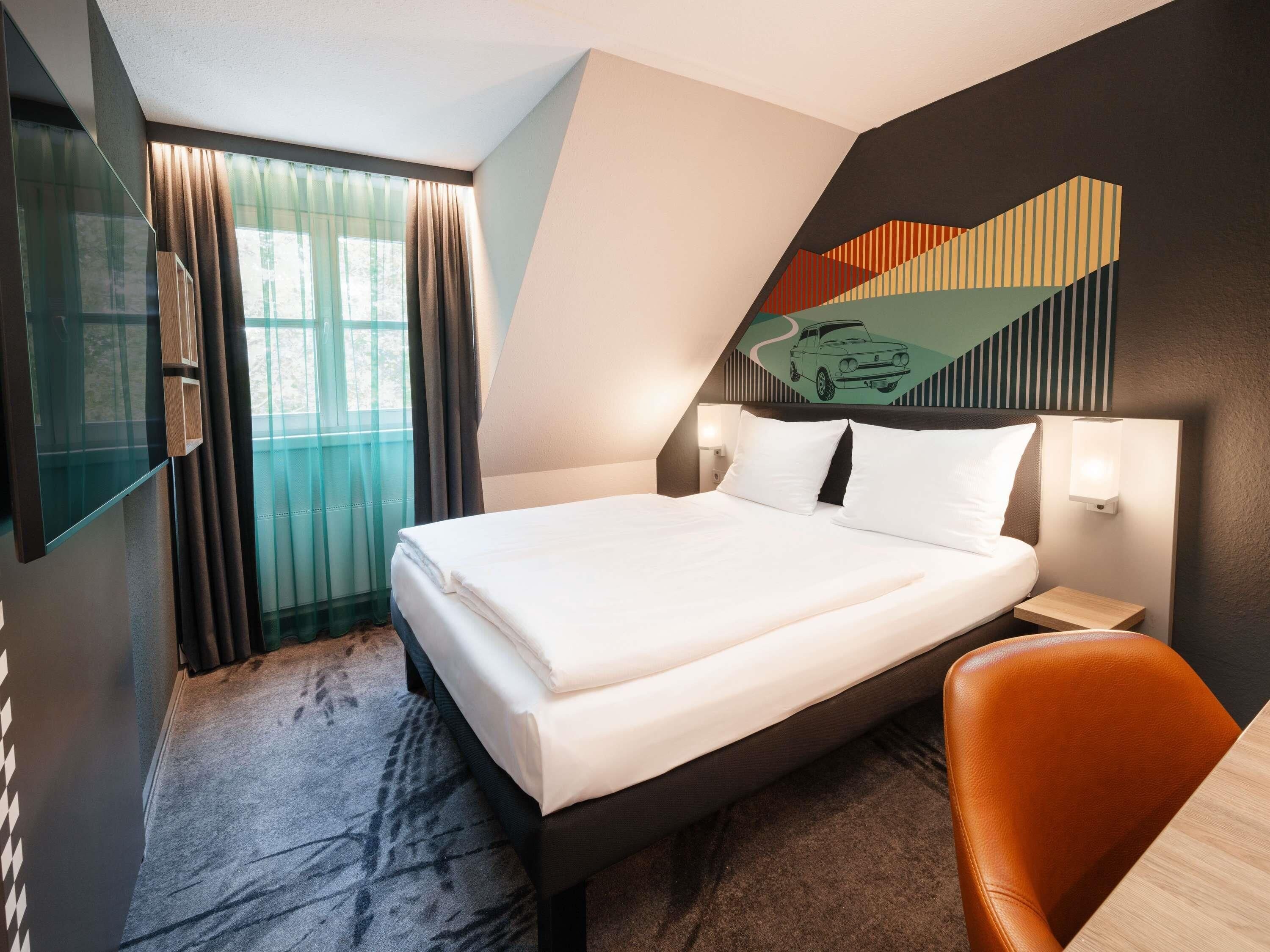 A modern hotel room with a large bed, a desk, a chair, and a large window with curtains.