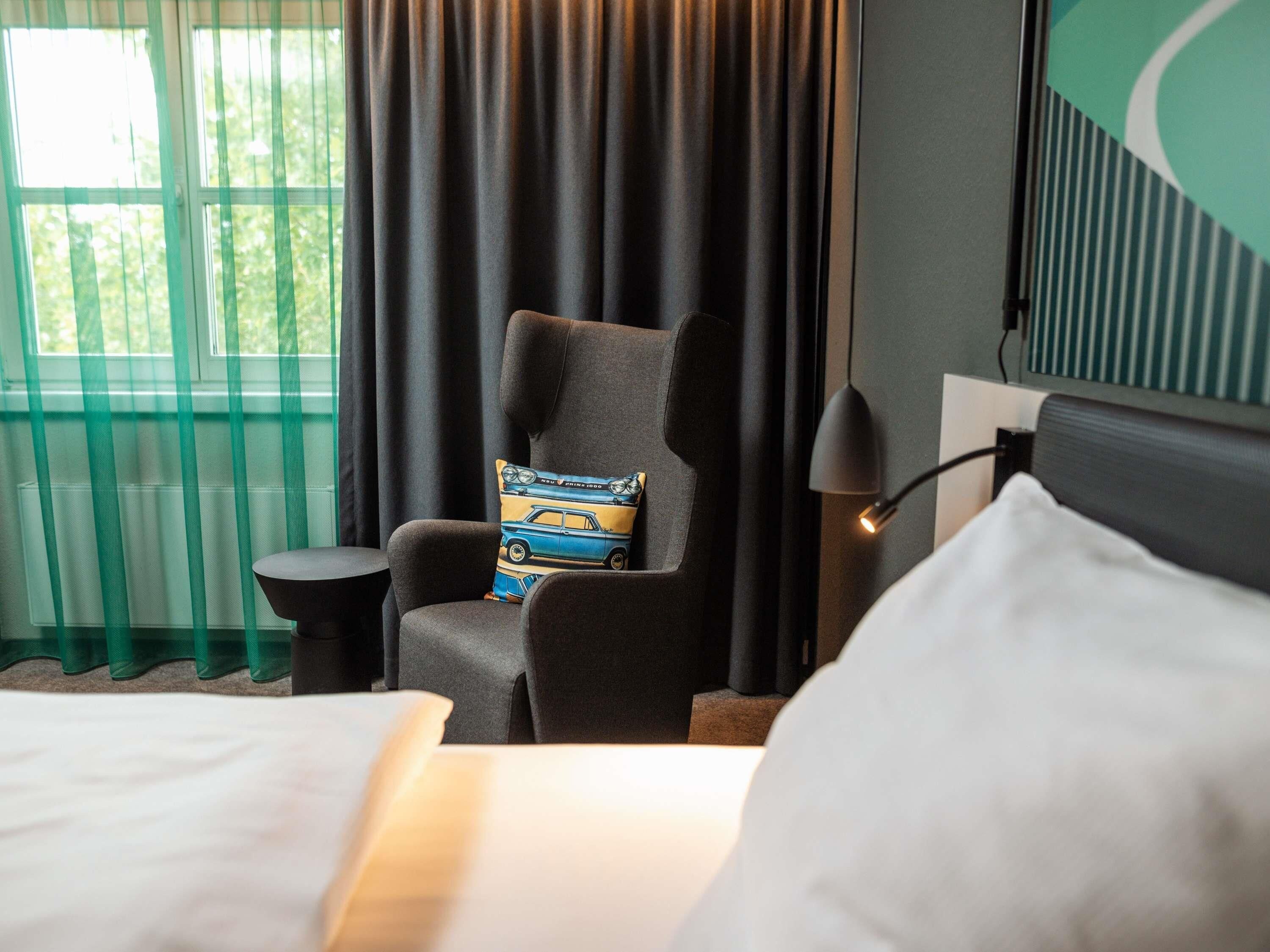 A hotel room with a bed, a chair with a car-themed pillow, a small side table, a lamp, and a view of greenery through the window.