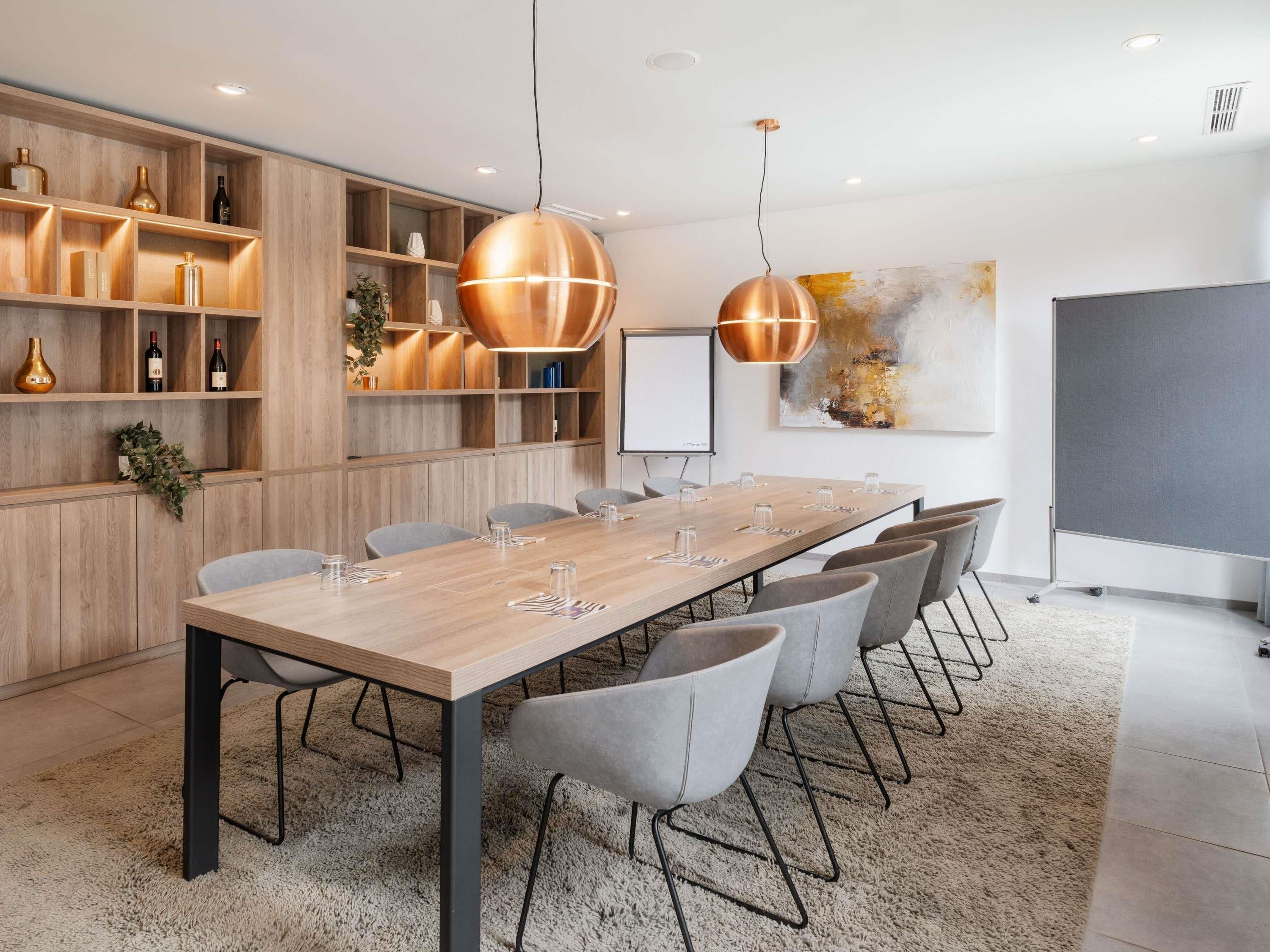 A conference room with a long table, grey chairs, wooden shelving, and two pendant lights.