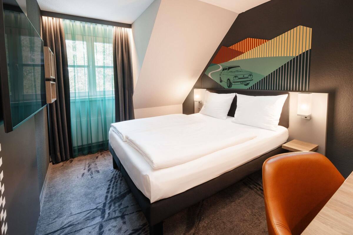 A modern hotel room with a large bed, a desk, a chair, and a large window with curtains.