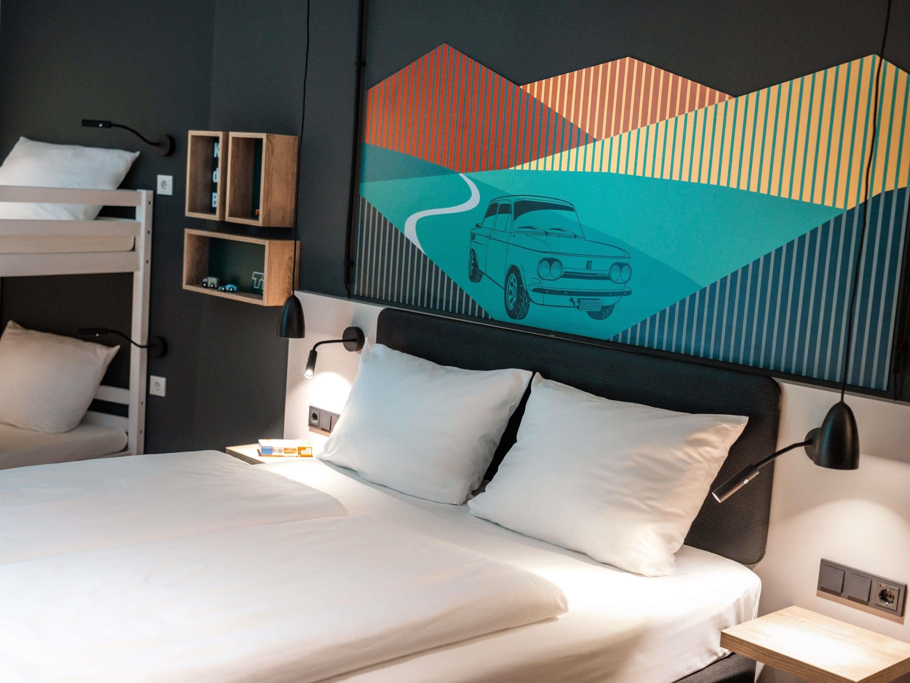 A modern hotel room with a bunk bed, a desk, a wall-mounted shelf, and a large abstract painting.