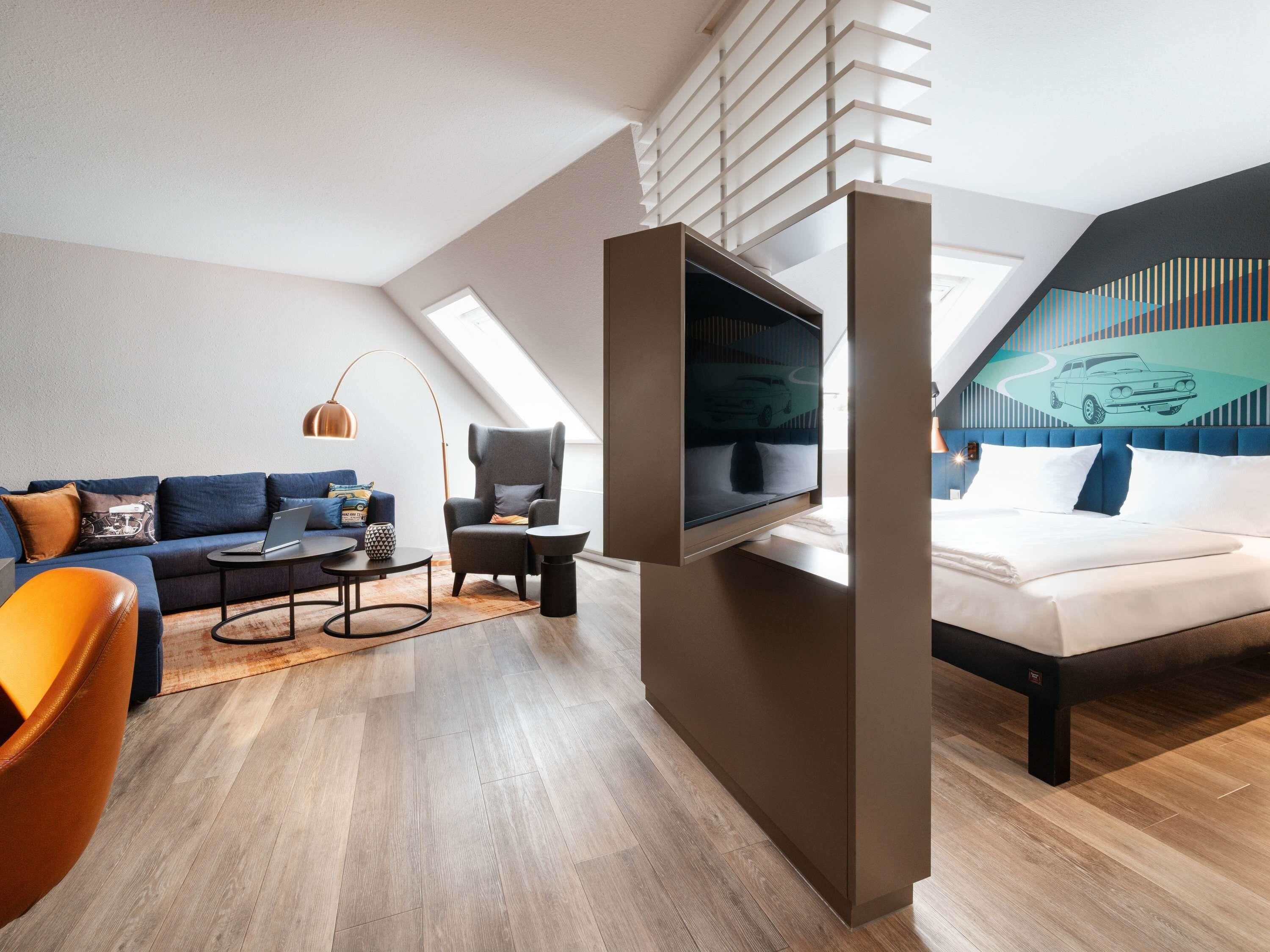 A modern loft bedroom with a large bed, a TV mounted on the wall, a sofa, a chair, and a desk.