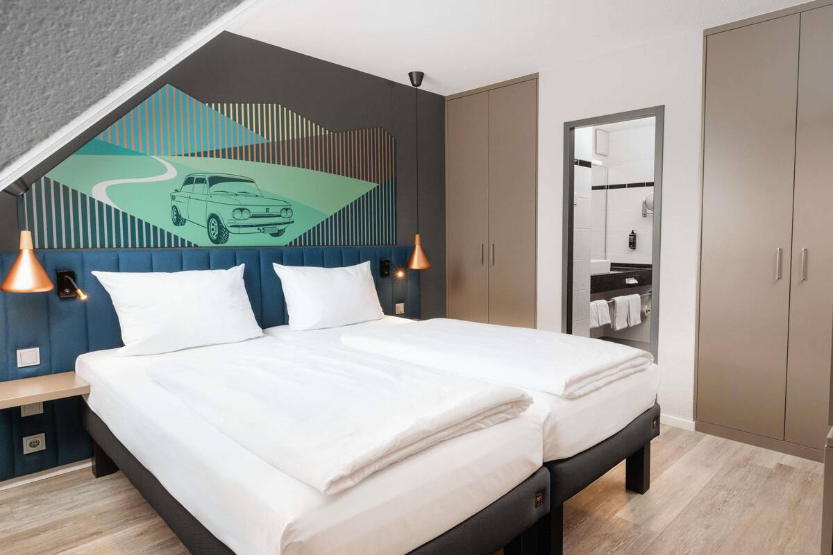 A modern hotel room with a bed, two bedside tables, a wall-mounted lamp, a wardrobe, and a bathroom mirror.