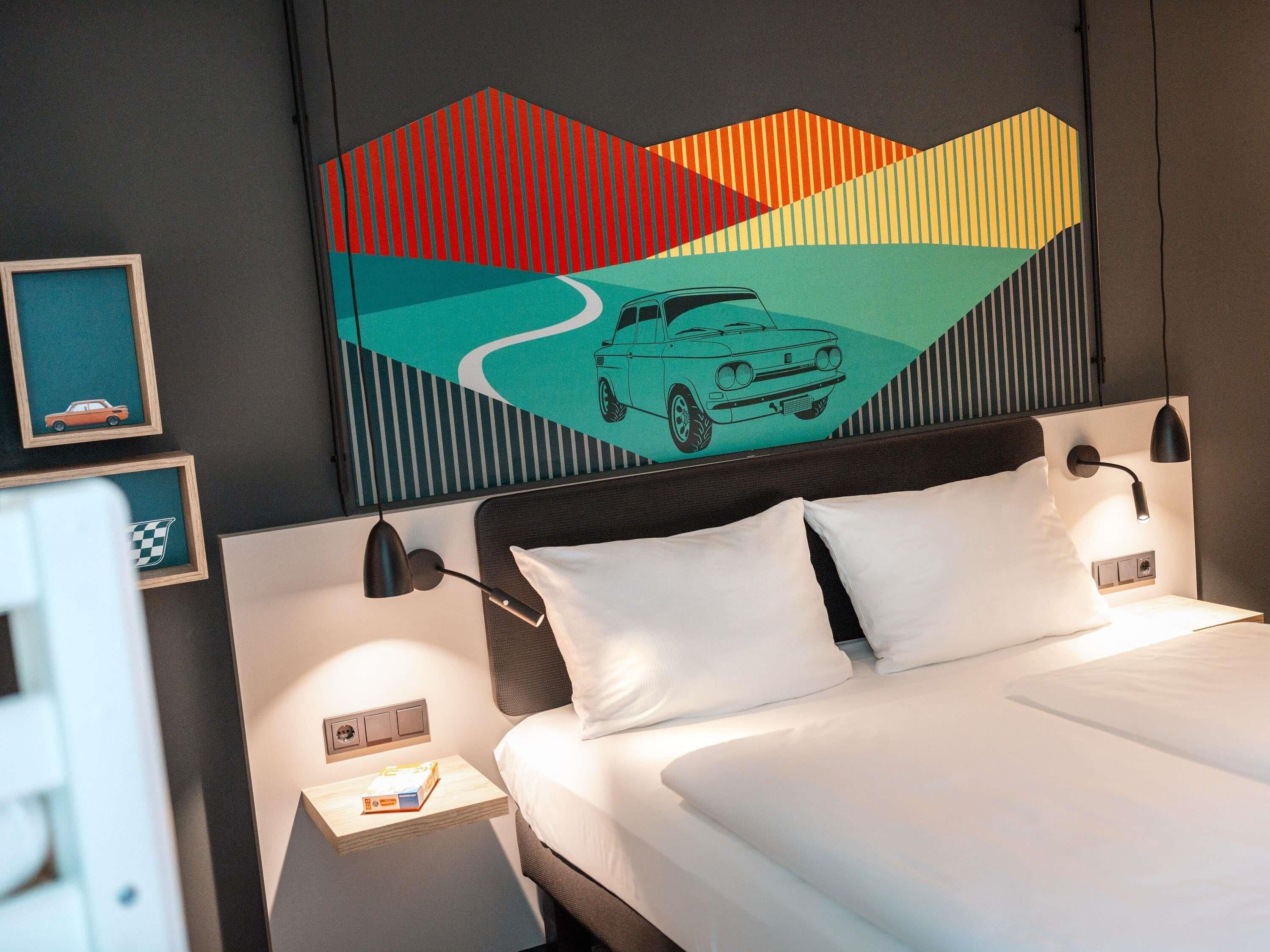Een hotelkamer met een bed, twee bedlampjes, een nachtkastje, een lijst met afbeelding en een wanddecoratie van een auto op een slingerende weg.