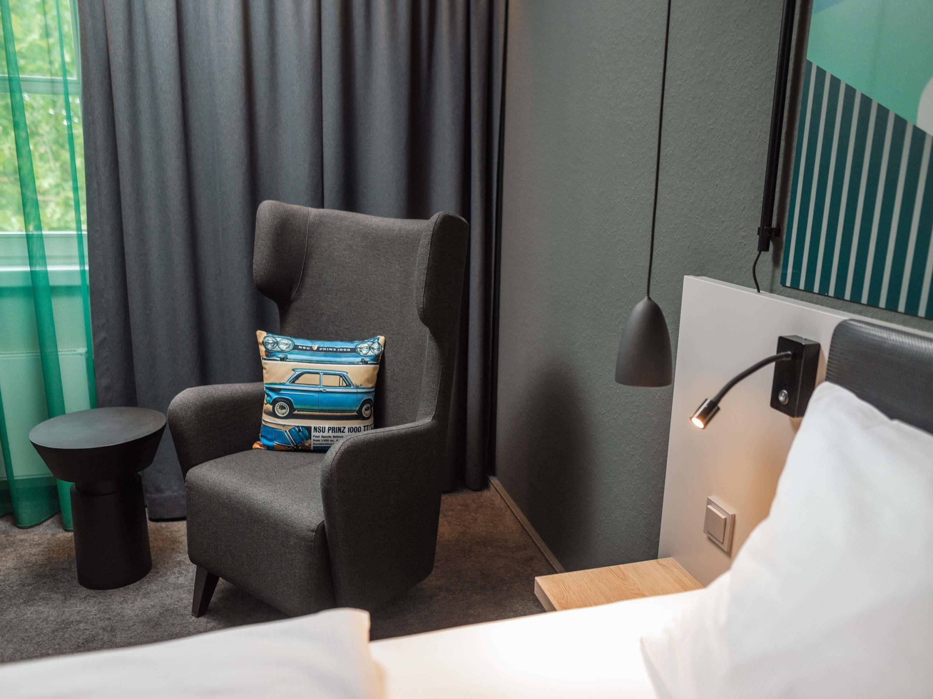 Une chambre d’hôtel moderne avec un fauteuil gris, une petite table ronde, une lampe de chevet et un lit recouvert de linge de maison blanc.