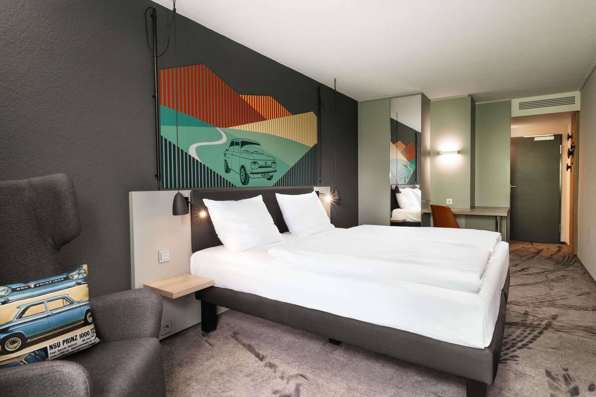 A hotel room with a large bed, a chair, a desk, and a wall with a geometric painting.