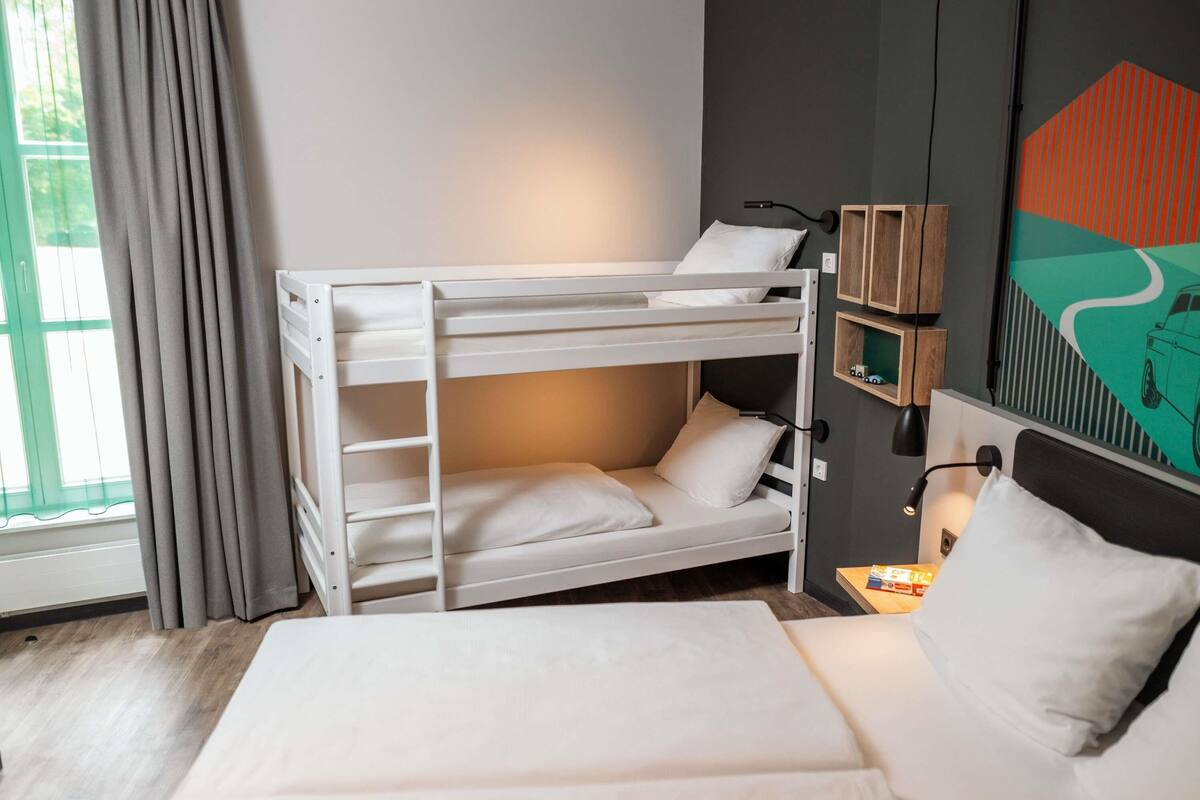 A bunk bed with a desk and chair, a bedside table with a lamp, a window with curtains, and a wall with a geometric painting.