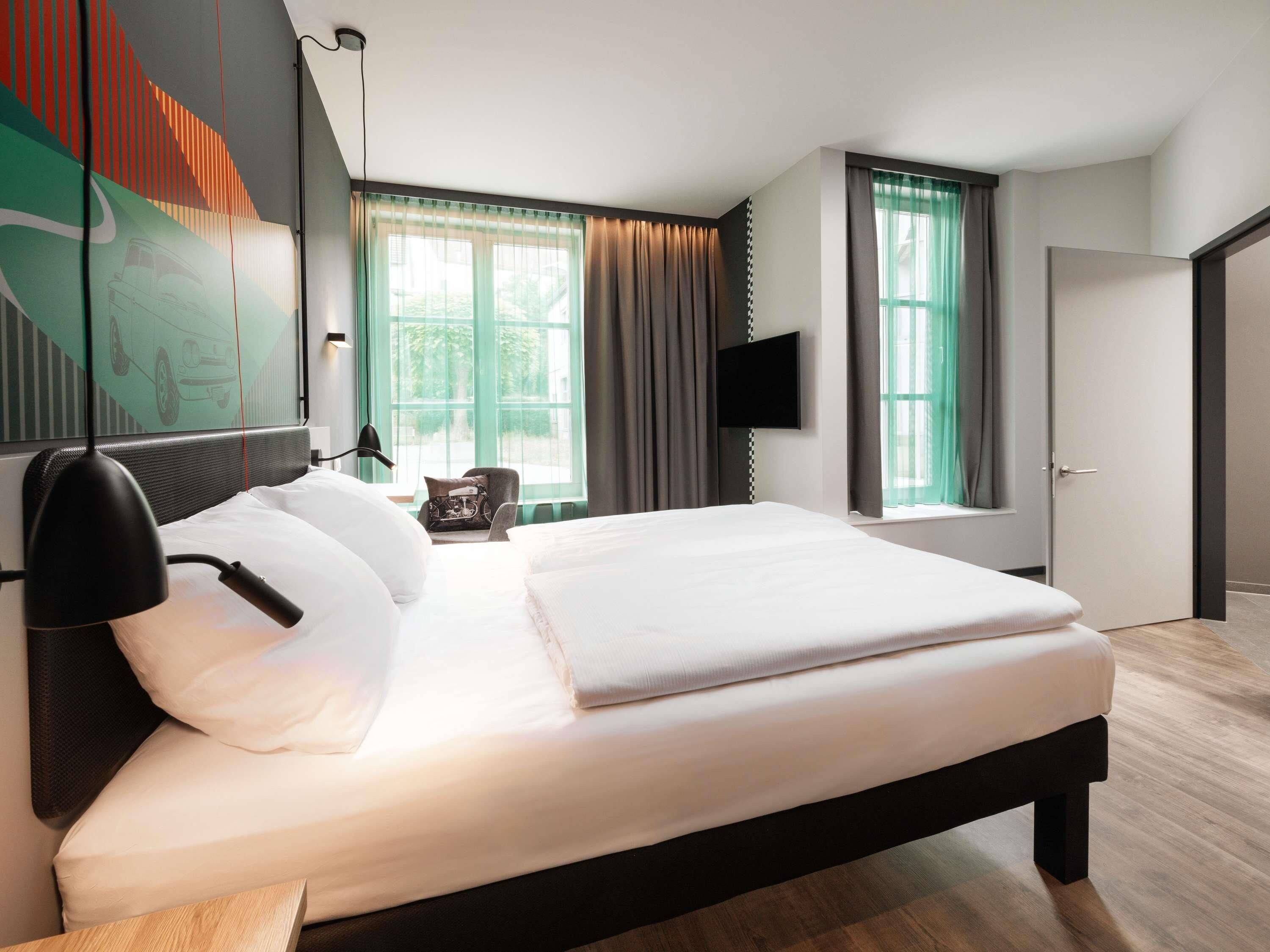 A modern hotel room with a large bed, a flat-screen TV, a chair, and a window with curtains.