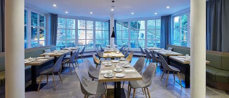 A spacious dining area with multiple tables and chairs, large windows offering a view of the outdoors, and a modern ceiling design with recessed lighting.