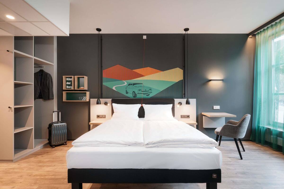 A hotel room with a bed, a desk, a chair, a wardrobe, and a wall with a geometric artwork.