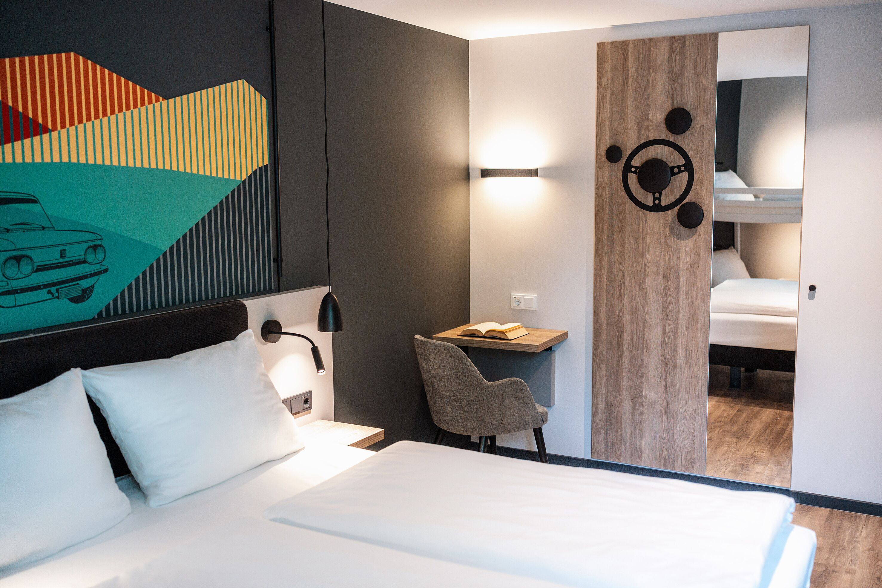A modern hotel room with a bed, a desk, and a chair. There is a painting on the wall and a mirror reflecting the room.