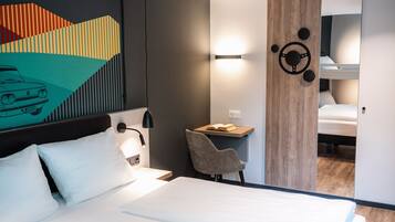 A modern hotel room with a bed, a desk, and a chair. There is a painting on the wall and a mirror reflecting the room.