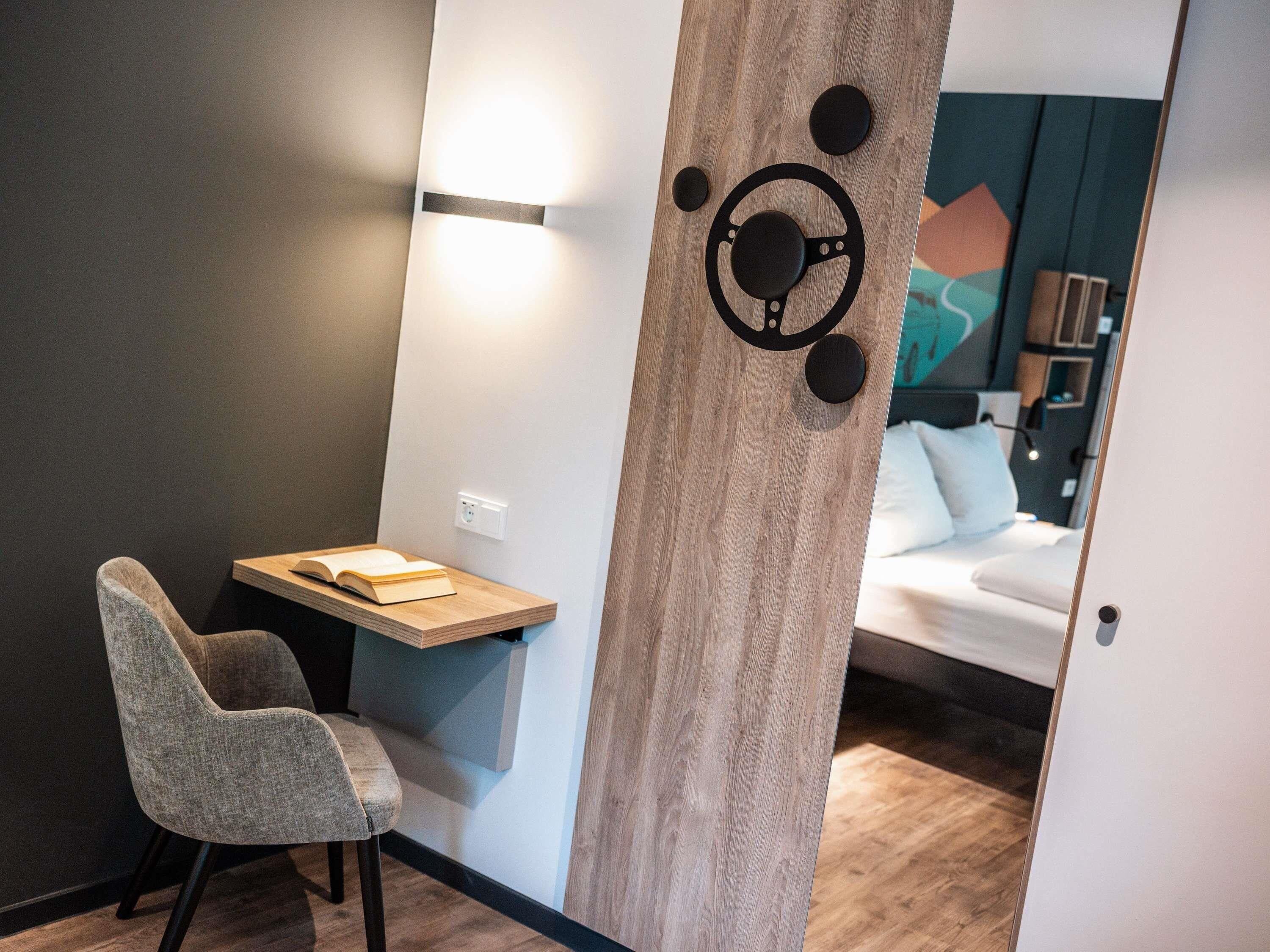 Ein modernes Hotelzimmer mit einem Holztisch, einem bequemen Sessel und einem Bett mit weißer Matratze und Kissen.