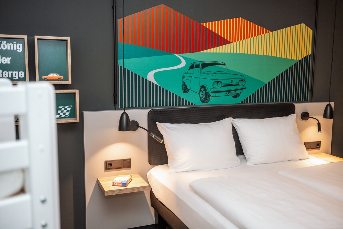 A hotel room with a bed, two bedside tables, a wall-mounted lamp, and a colorful abstract painting.