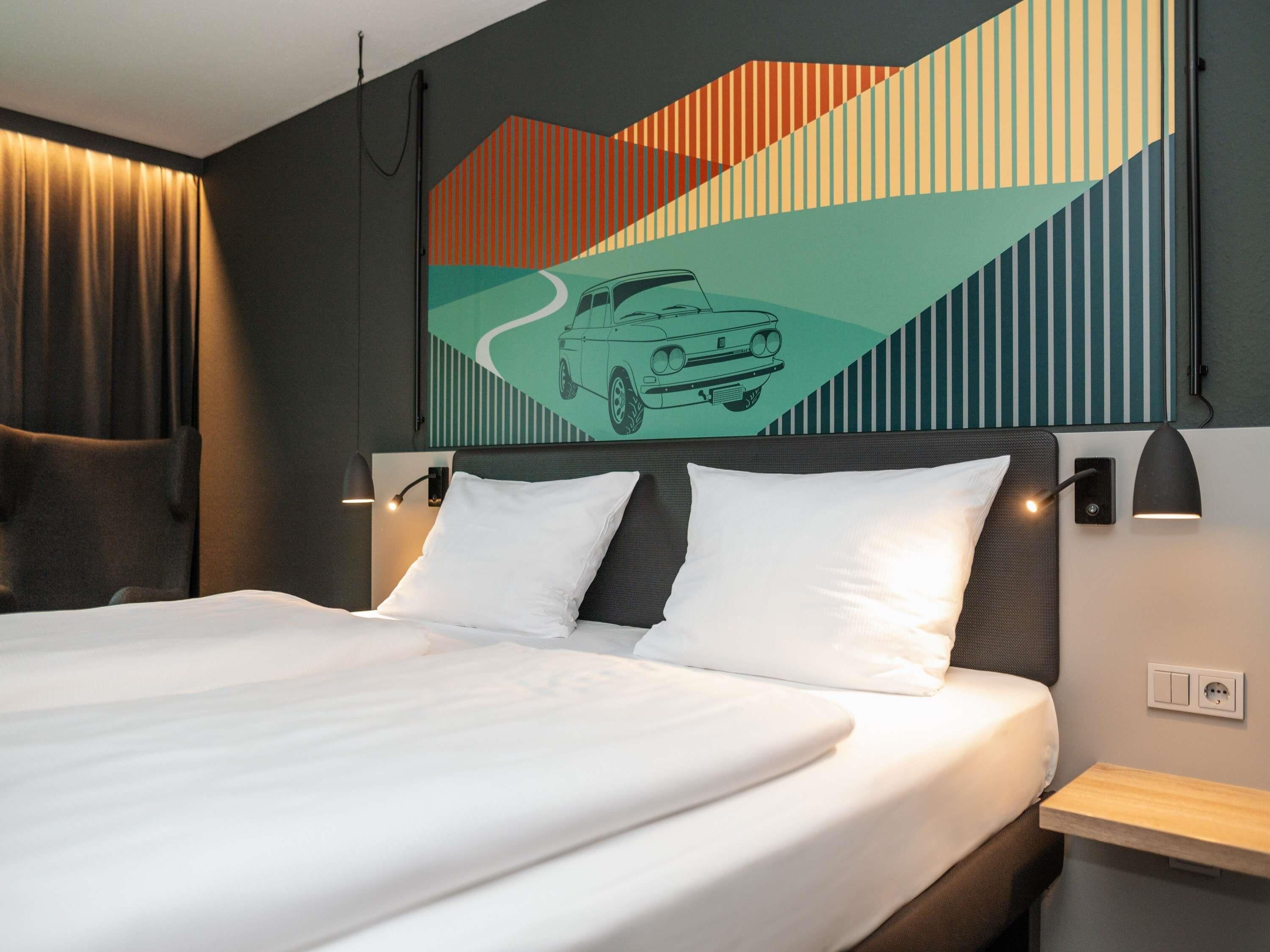 Quarto de hotel com uma cama grande, duas mesas de cabeceira, cabeceira com ilustração de carro e uma luminária fixada na parede.